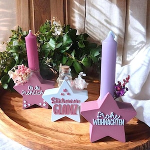 May include: A wooden tray holds three star-shaped candle holders with candles. The candle holders are pink and white, with text that reads "Oh du Fröhlich", "Sternen GLANZ", and "Frohe Weihnachten". The candles are pink and purple.
