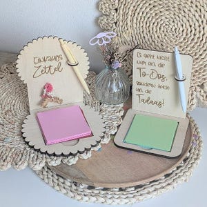 May include: Two light-colored wooden note holders with scalloped or square edges. One features the text 'Einkaufs Zettel' with pink sticky notes and a pen. The other features the text 'Es geht nicht nur um die To-Dos, sondern auch um die Tadaas!' with green sticky notes and a white pen.
