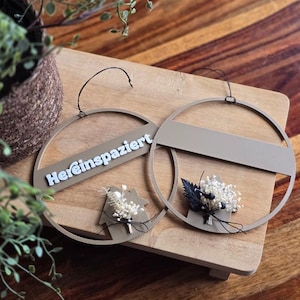 Puede incluir: Dos adornos de pared circulares de color topo con detalles florales y la palabra "Hereinspaziert" en blanco. Uno presenta un recorte en forma de casa con flores secas, el otro un ramo. Se exhiben sobre una superficie de madera.