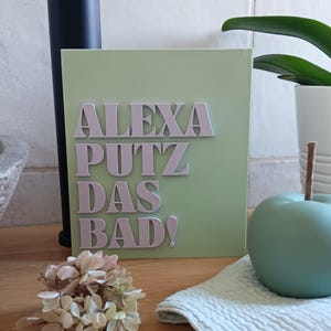 Op de afbeelding: Een lichtgroen vierkant bord met de tekst "Alexa putz das bad!" in witte letters. Het bord staat op een houten oppervlak.