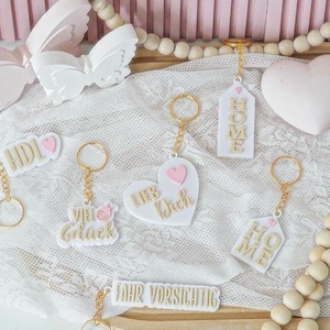 Könnte beinhalten: Eine Sammlung weißer Schlüsselanhänger mit goldfarbenen Schlüsselringen. Einige Schlüsselanhänger sind herzförmig mit einem rosa Herz und den Worten "Lieb Dich". Andere Schlüsselanhänger haben das Wort "HOME" oder "VIEL Gluck".