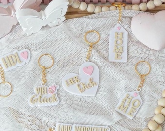 Peut inclure: Plusieurs porte-clés blancs avec des anneaux dorés. Certains sont en forme de cœur avec un cœur rose et l'inscription "Lieb Dich". D'autres portent les mots "HOME" ou "VIEL Gluck".