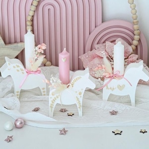 May include: White wooden horse candle holders with gold detailing, holding white and pink candles. The candle holders are decorated with dried flowers and pink ribbons. Decorative stars and spheres are scattered on the white lace surface.