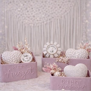 Puede incluir: Tres pequeños recipientes rectangulares, de color púrpura claro, con objetos blancos en forma de corazón y flores secas. Los recipientes tienen texto en relieve en alemán. Un recipiente tiene un pequeño objeto redondo con texto.