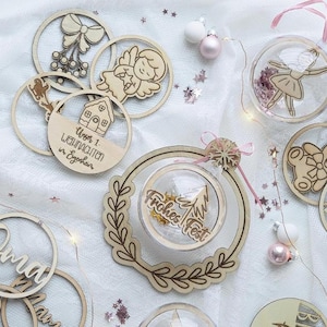 May include: A collection of wooden Christmas ornaments and clear baubles. The wooden ornaments feature cut-out designs of a bow, angel, house, and text. The baubles contain a ballerina and a Christmas tree with the text "Frohes Fest".