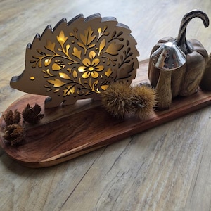 May include: A wooden hedgehog-shaped decorative light with floral cutouts, illuminated with warm light, sits on a wooden tray. Accompanying the hedgehog are decorative wooden mushrooms, pinecones, and a pumpkin. The tray is made of dark brown wood.