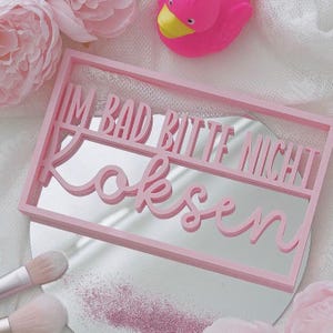 Op de afbeelding: Een roze rechthoekige spiegel met de tekst "IM BAD BITTE NICHT Koksen" in cursieve letters uitgesneden. De spiegel is omgeven door roze bloemen, make-up borstels, een roze badeend en roze glitter.
