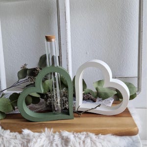 Pode incluir: Dois vasos em forma de coração, um verde sálvia e outro branco, estão numa superfície de madeira. O vaso verde contém um tubo de vidro com uma rolha de cortiça. Eucalipto e outras plantas verdes estão dispostas ao redor.