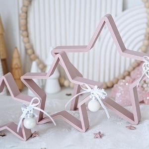 May include: Three light pink wooden star decorations with white ornaments and ribbons. The stars are arranged on a white surface with small star-shaped confetti. The background includes a beaded wreath and other decorative elements.