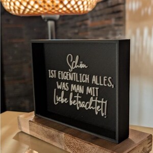 Puede incluir: Un letrero rectangular negro con el texto alemán "Schön ist eigentlich alles, was man mit Liebe betrachtet!" en blanco. El letrero está montado sobre una base de madera.