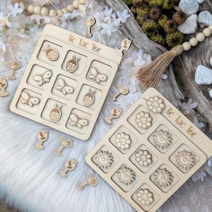 May include: Two wooden Tic Tac Toe game boards with laser-cut designs. One board features ladybirds, butterflies, and cocktail glasses. The other board has flowers and suns. The words "Tic Tac Toe" are engraved on each board.