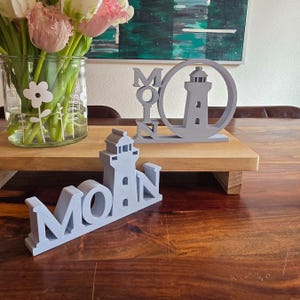 Puede incluir: Dos letreros de madera grises con la palabra "Moin" y un diseño de faro. Los letreros están sobre una mesa de madera con un jarrón de flores en el fondo.