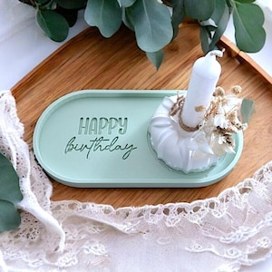 Op de afbeelding: Een lichtgroene ovale schaal met de woorden "Happy Birthday" erop gegraveerd. Een witte kaarsenhouder met een witte kaars en gedroogde bloemen staat op de schaal.