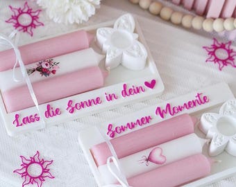 May include: Two white rectangular trays with candles. Each tray holds three candles: two pink and one white. The white candles have floral or heart designs. The trays have text in pink. White ribbons and flower-shaped candle holders are also present.