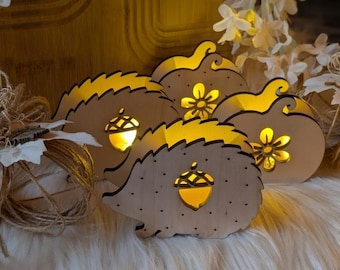 May include: Wooden decorative lights in the shape of hedgehogs and pumpkins. The lights have a warm glow from within, with acorn and flower cutouts. The lights are arranged on a fluffy white surface, with decorative foliage in the background.