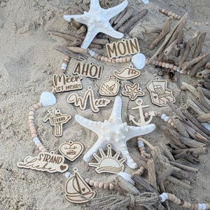 Può includere: Ornamenti in legno a tema spiaggia e stelle marine disposti sulla sabbia. Gli ornamenti includono disegni nautici come un'ancora, una barca a vela e stelle marine, insieme a frasi come "MOIN" e "Strand Liebe".