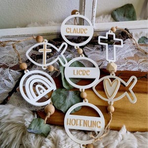 Può includere: Ornamenti in legno bianchi con simboli religiosi e parole. Gli ornamenti includono una croce, un pesce, un arcobaleno e cerchi con le parole "CLAUDE", "LIEBE" e "HOFFNUNG". Gli ornamenti sono collegati con perline di legno e spago.