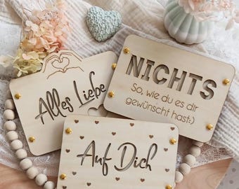 May include: Three wooden signs with different messages. The first sign says "Alles Liebe" with a heart symbol. The second sign says "Nichts So, wie du es dir gewünscht hast:". The third sign says "Auf Dich" with small hearts surrounding the text.