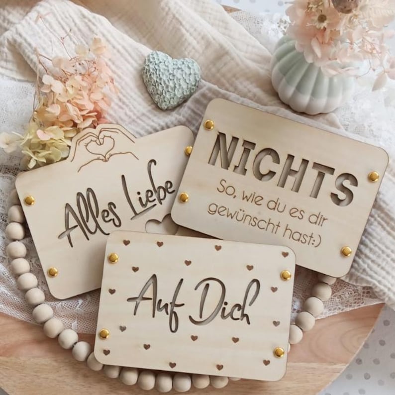 May include: Three wooden signs with different messages. The first sign says "Alles Liebe" with a heart symbol. The second sign says "Nichts So, wie du es dir gew&uuml;nscht hast:". The third sign says "Auf Dich" with small hearts surrounding the text.