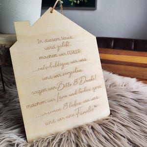 May include: A wooden house-shaped sign with engraved text in German. The sign has a natural wood finish and a leather cord for hanging. The text includes phrases like "In diesem Haus wird gelebt" and "sind wir eine Familie."