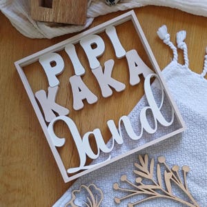 Könnte beinhalten: Holzschild mit den Worten "PIPI KAKA Land" in weißen und beigen Buchstaben. Das Schild liegt auf einer Holzoberfläche mit einem weißen Tuch und dekorativen Holzausschnitten im Hintergrund.