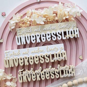 May include: Three decorative signs with the German phrase "Einfach mal machen, könnte ja UNVERGESSLICH werden" in a layered design. The signs are in white and beige, with a pink rainbow background and floral accents.