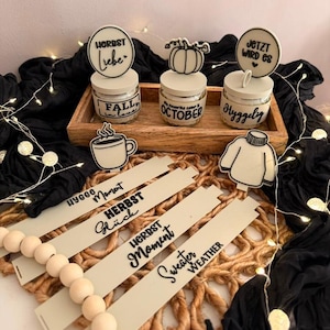 May include: Three glass jars with cream-coloured lids and circular toppers featuring text and pumpkin graphics, placed in a wooden tray. Includes flat, cream-coloured wooden tags with black text reading Hygge Moment, Herbst Glück, Herbst Moment, and Sweater Weather.