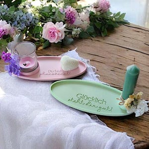 May include: Two oval-shaped decorative trays with inspirational messages. The pink tray has a heart-shaped object and a glass candle holder. The green tray has a candle and a small bouquet of dried flowers. The text on the pink tray reads "Du bist phenomenal!" and the text on the green tray reads "Gl&uuml;cklich steht dir gut!"