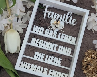 May include: A white framed sign with the German word "Familie" at the top, followed by text in German. A heart-shaped decoration with gold accents is in the center. The sign is surrounded by white flowers and greenery.