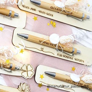 May include: Several bamboo pens with silver detailing are presented on wooden tags. The tags feature German text and decorative elements. The background is a soft pink with scattered star-shaped confetti. The tags have phrases like "Neues Jahr - Neues Glück".