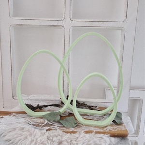 May include: Three light green egg-shaped cookie cutters. The cutters are arranged on a wooden board with a white lace and faux fur background. The cutters are made of plastic and are a pastel green color. The cutters are for baking.