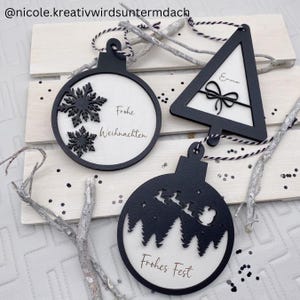 May include: Black and white Christmas tree decorations in various shapes. One round ornament features snowflakes and the text "Frohe Weihnachten." Another round ornament shows a silhouette of Santa's sleigh and the text "Frohes Fest." A third ornament is a triangle with the name "Emma."