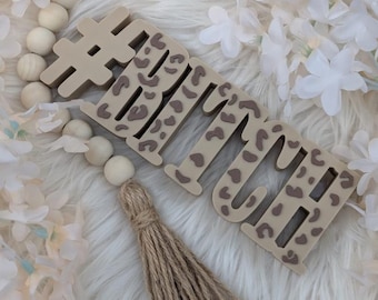 May include: A wooden sign with the text "#BITCH" in brown and tan leopard print. The sign is hanging from a wooden bead garland with a brown tassel.