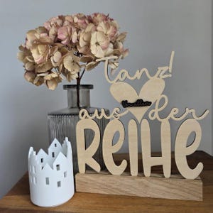 Könnte beinhalten: Ein dekoratives Holzschild mit dem deutschen Satz "Tanz aus der Reihe" auf einem Holzsockel. Ein weißer Keramik-Kerzenhalter in Hausform und eine Glasvase mit getrockneten Blumen sind ebenfalls im Bild.