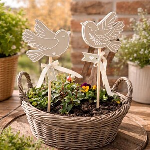 May include: Two decorative bird stakes in a wicker basket filled with plants. The birds are light brown with white outlines and are attached to wooden sticks. Each bird has a white ribbon and a small banner with a heart. The basket is on a wooden surface.