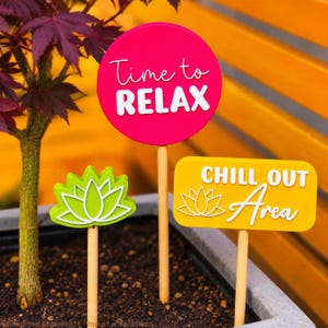 May include: Three garden stakes with wooden dowels. One round hot pink sign reads Time to RELAX in white text. One rectangular yellow sign reads CHILL OUT Area with a white lotus icon. One green lotus-shaped sign with white outline details.