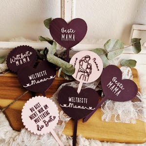 Puede incluir: Una colección de adornos decorativos en varias formas y colores. Los adornos incluyen formas de corazón y circulares en tonos burdeos y rosa, con texto blanco que dice "Beste Mama" y "Weltbeste Mama". Algunos adornos presentan una ilustración de madre e hijo.