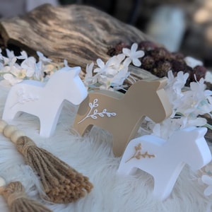 May include: Three wooden horse figurines with a white and brown paint finish. Each figurine has a white floral design painted on the side. The figurines are sitting on a white fluffy surface.