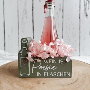 Puede incluir: Un soporte de madera para botellas de color verde oliva con un dibujo lineal blanco de una botella de vino con un corazón y el texto blanco "WEIN IST Poesie IN FLASCHEN". Una botella de vidrio transparente con líquido rosa está rodeada de flores.