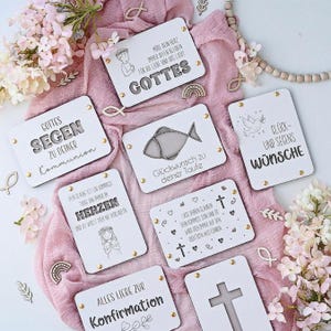 Könnte beinhalten: Eine Sammlung weißer Holztafeln mit religiösen Themen, die gravierten Text und Illustrationen aufweisen. Die Tafeln enthalten Sätze wie "GOTTES" und "Konfirmation" mit dekorativen goldfarbenen Akzenten. Die Anordnung befindet sich auf einem rosa Stoff.