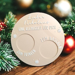 Op de afbeelding: Een beige, ronde houten kerstversiering met twee cirkelvormige uitsparingen en witte tekst. De tekst bevat "Lieber Nikolaus" en "Eine Kleinigkeit für Dich". Er zijn ook sneeuwvlokontwerpen.