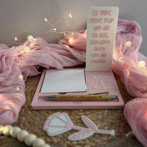 Peut inclure: Un organisateur de bureau rose clair comprenant un panneau vertical avec le texte allemand Es geht nicht nur um die To-dos, sondern auch um die Tadaas!, un bloc-notes carré, un stylo marqué Hokus Pokus et une petite découpe en forme de fleur.