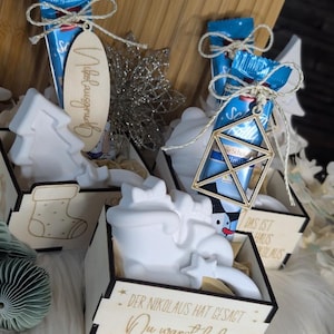 May include: Wooden boxes with Christmas-themed decorations, including a tree, boot, and Santa Claus. Each box contains a blue chocolate bar tied with twine and a wooden tag. The boxes are arranged on a wooden surface.