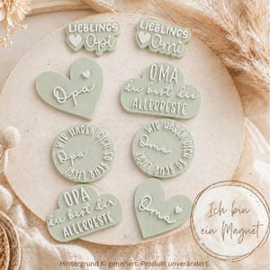 May include: A selection of sage green magnets with white text. The magnets are in various shapes, including hearts and circles, and feature phrases such as "Lieblings Opi" and "Oma du bist die ALLERBESTE". The text "Ich bin ein Magnet" is also visible.