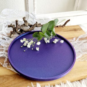 以下が含まれることがあります： わずかに盛り上がった縁のある丸い紫色のトレイ。トレイは滑らかでマットな素材でできており、微妙なグラデーション効果があります。トレイは白い花と緑の葉の枝で飾られています。