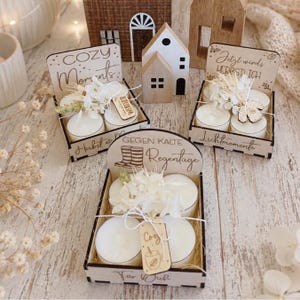 May include: Three wooden gift boxes containing white tea light candles, dried white flowers, and small wooden tags. The boxes feature laser-engraved text including Cozy Moments, Herbst, Jetzt wirds herbstlich, Lichtmomente, and Gegen kalte Regentage.