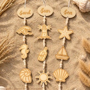 May include: Three vertical hanging decorations made of light-coloured wood beads and laser-cut shapes. The top ovals feature the words sand, sun, and sea. The hanging ornaments include a wave, lighthouse, starfish, seahorse, message in a bottle, sailboat, spiral shell, sun, and scallop shell.