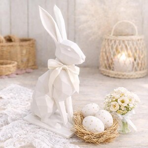 Könnte beinhalten: Eine weiße, geometrische Osterhasenfigur mit cremefarbener Schleife. Daneben ein Nest mit drei Eiern, die mit goldenen Punkten verziert sind, und ein kleiner Strauß weißer Blumen. Im Hintergrund eine dekorative Laterne.