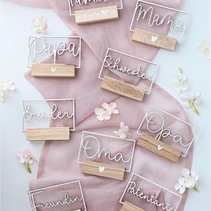 Könnte beinhalten: Eine Sammlung personalisierter Namenskarten mit weißer Drahtschrift auf klarem Acryl, platziert auf kleinen Holzblöcken. Die Namen umfassen "Papa", "Mama" und andere, mit einem kleinen Herzdetail auf jedem Block. Die Karten sind auf einem rosa Stoff angeordnet.