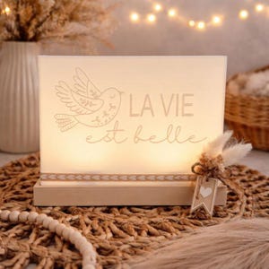 Könnte beinhalten: Ein rechteckiges, beleuchtetes Schild mit dem Schriftzug "LA VIE est belle" und einer Vogelillustration. Das Schild steht auf einem Holzsockel und hat ein dekoratives Etikett mit einem Herz. Das Design wirkt sanft und warm.